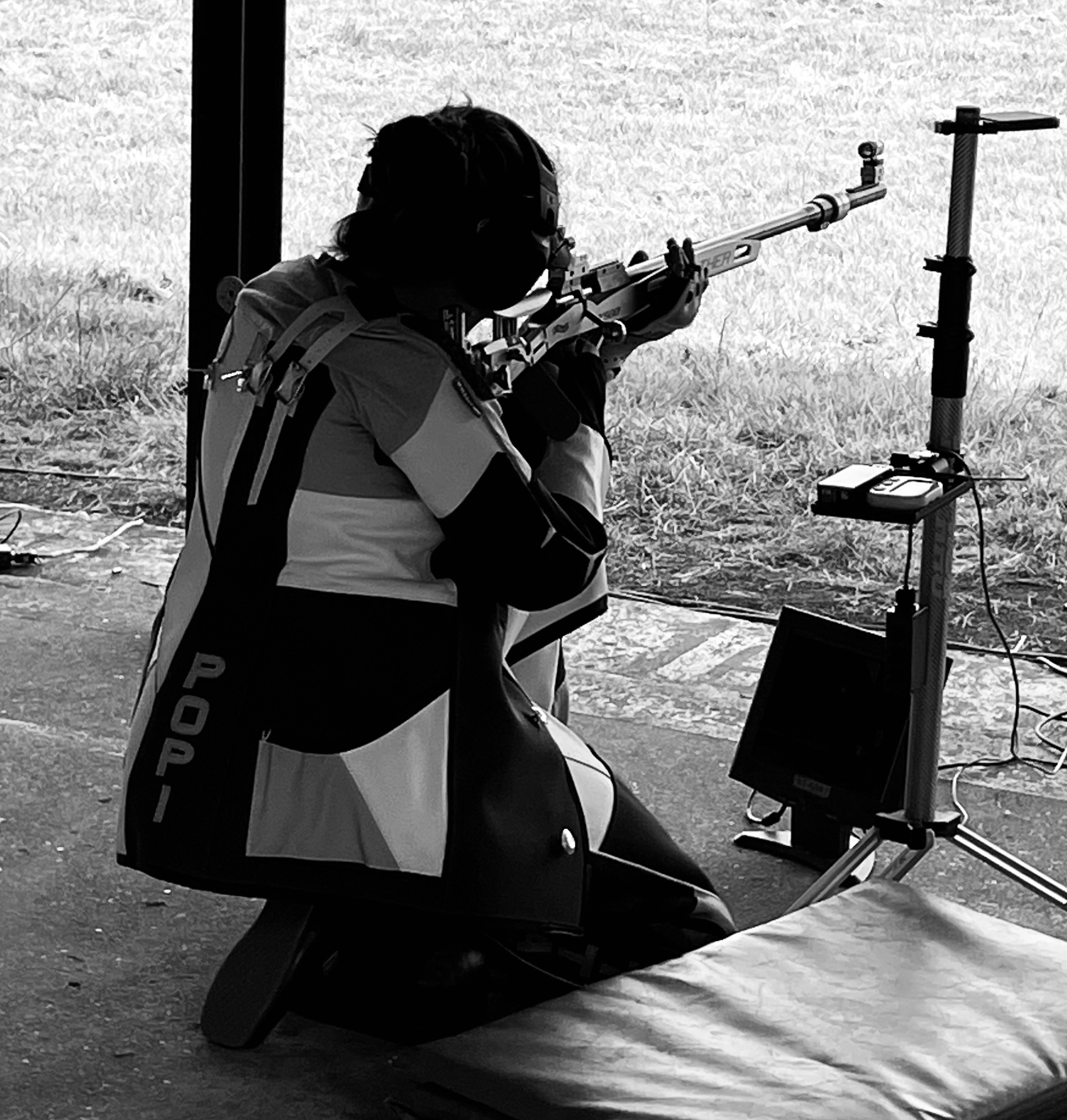 Popi Settas kneels faced away from the camera firing a rifle in a competition