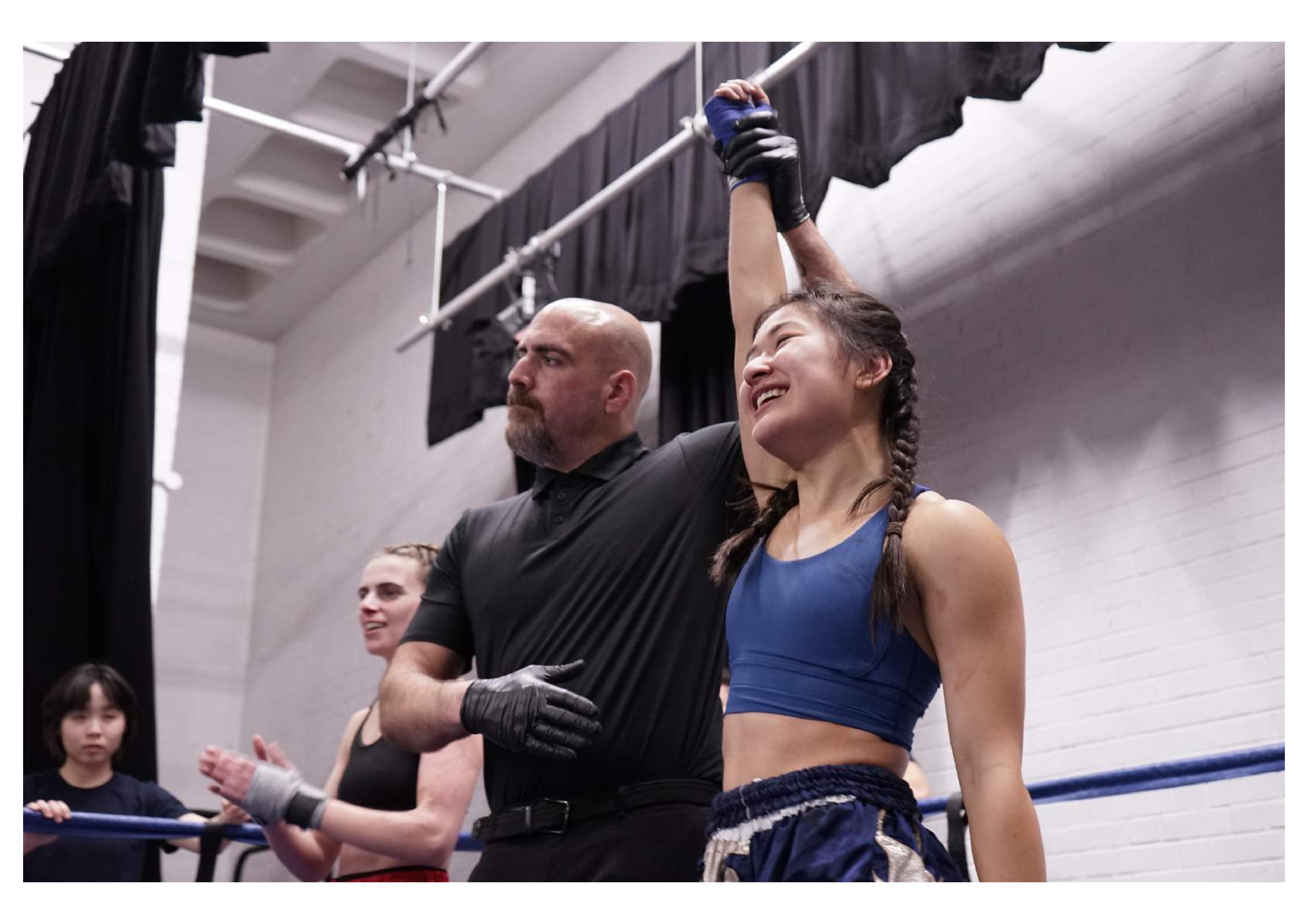 Female fighter wearing blue with hand raised in a boxing ring after a fight next to referee and opponent