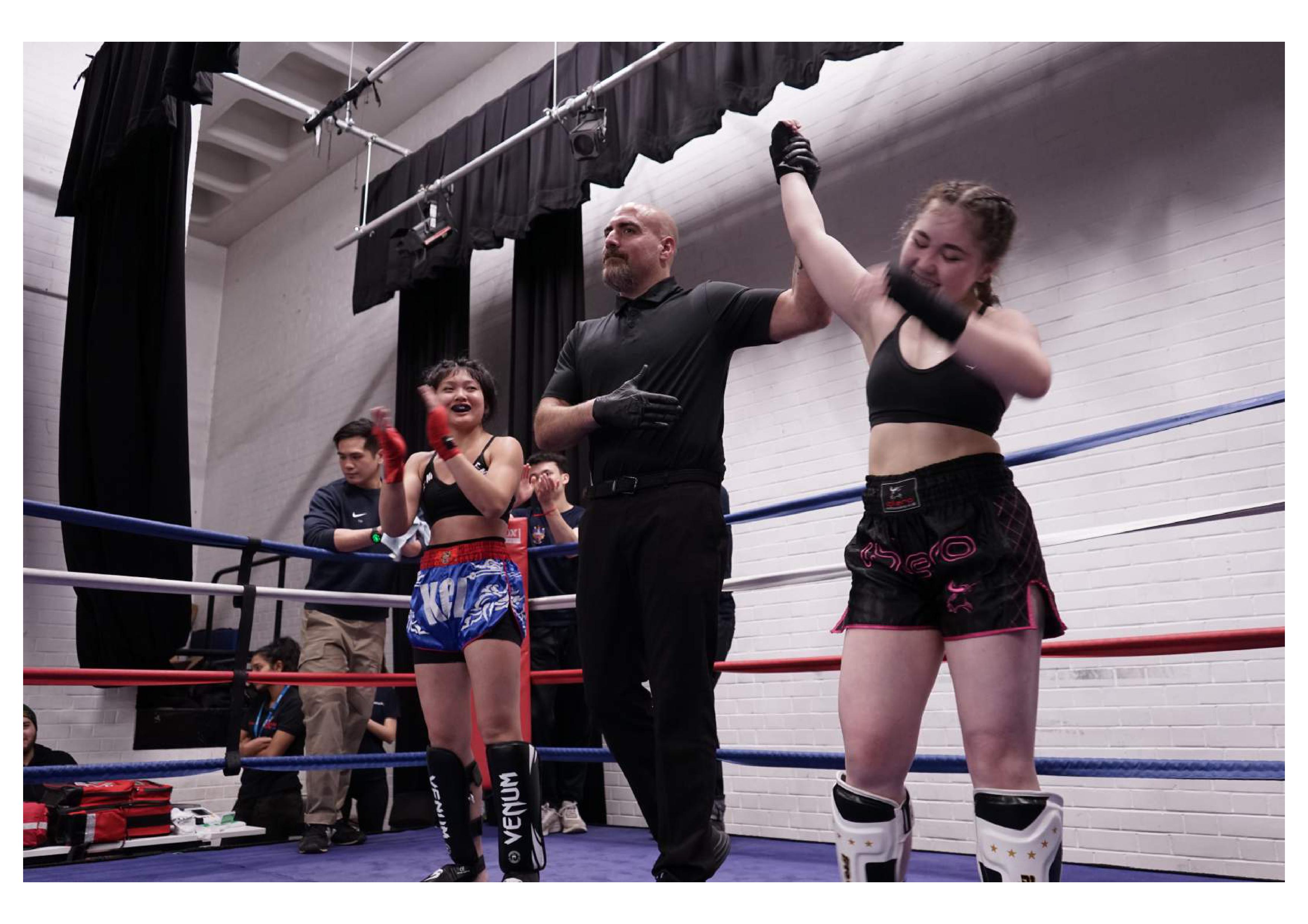 Female fighter wearing black with hand raised in a boxing ring after a fight next to referee and opponent