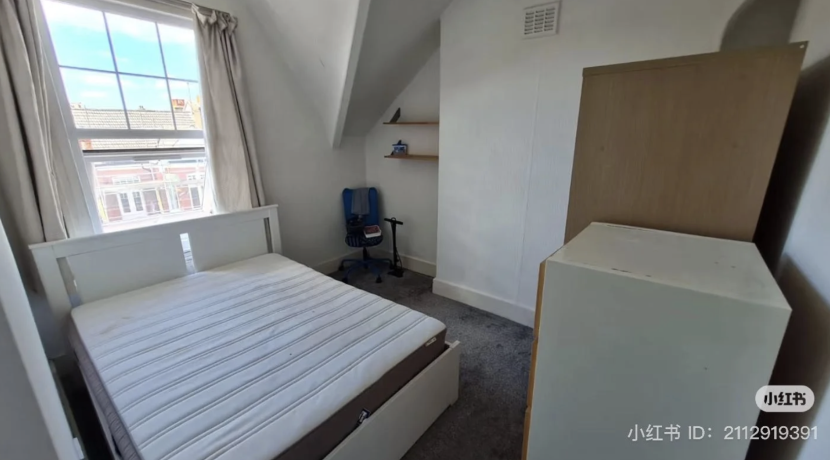 A bright, compact bedroom with a large window letting in natural light. The room features a double bed with a white frame and mattress, a blue desk chair with a few items on it, two wooden wall shelves, a wardrobe, and a white chest of drawers. The room has sloped ceilings and grey carpet flooring, creating a cozy and clean atmosphere. Ideal for students or young professionals.