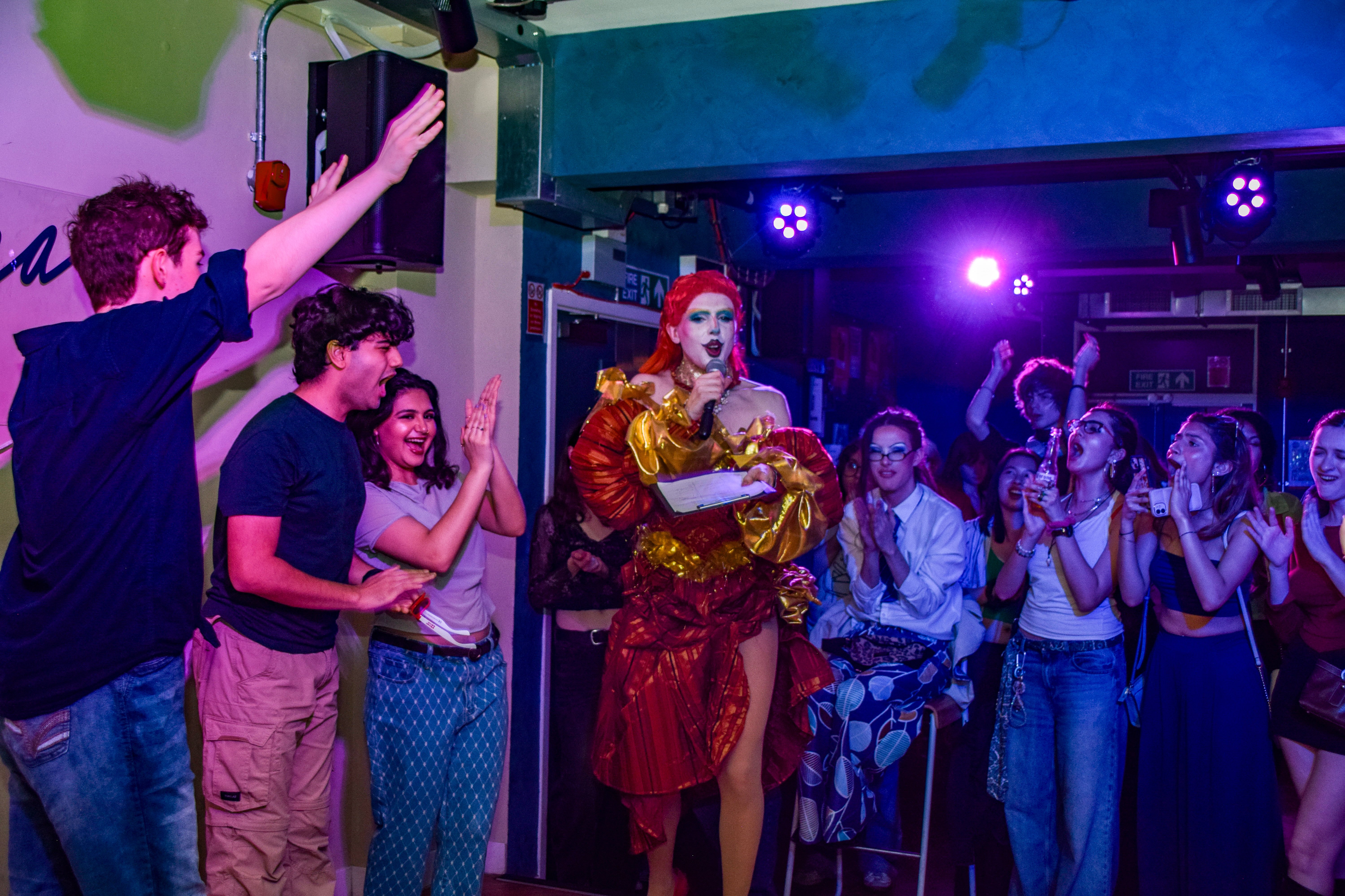 Drag Queen Bonk stands on stage talking with students who are all clapping and laughing