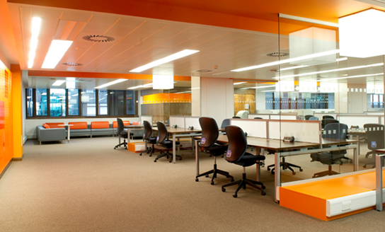 A bright modern space with glass partitions and orange walls, with soft seating as well as desk chairs