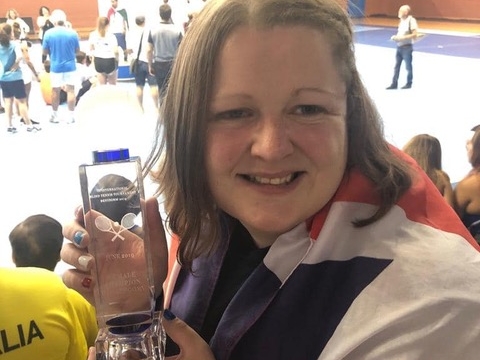 Image of Rachel from the shoulders up holding a tennis trophy. The trophy is clear. She is wrapped in a United Kingdom flag and her nails are painted red, white and blue to match.