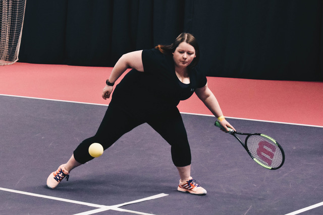 Rachel (author) playing tennis. Full body shot holding a tennis racket. Rachel is wearing all black and has long straight brown hair and pink trainers.