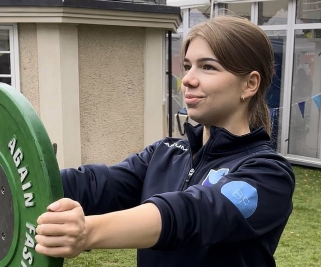 This photo shoes Natalia holding a green weightlifting plate. She is looking off to the left, and is smiling. Natalia is Caucasian, has light brown hair and dark eyes. 