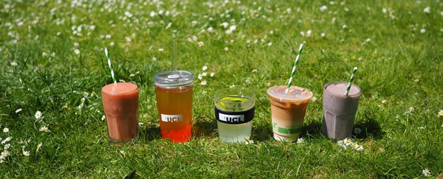Row of cold drinks lined up on grass on a summer day. From L-R: Strawberry smoothie in a glass, passionfruit bubble tea in a UCL reusable cup, lemon iced tea in a UCL reusable glass cup, Iced latte in a disposable cup and berry blast protein milkshake in a glass.